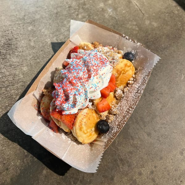 Mini pancakes with whipped cream, red and blue sprinkles, strawberries, and blueberries in a paper tray.