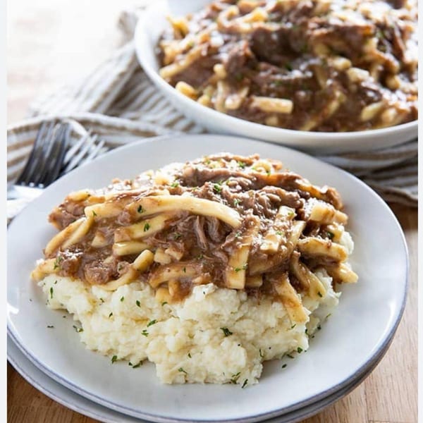 Hearty beef and noodles with gravy over mashed potatoes on a white plate, with a bowl in the background.