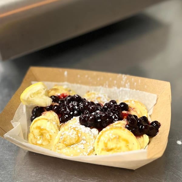 Mini pancakes topped with blueberries, powdered sugar, and a lemon wedge in a paper tray.